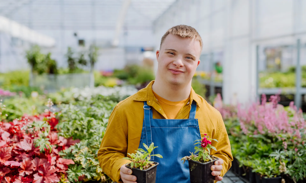 IMAGE DESCRIPTION: A Client wearing an apron is standing in a greenhouse holding two potted seedlings