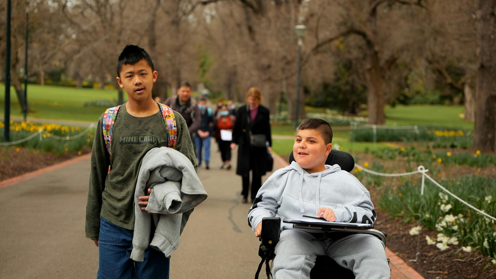Image of two school aged males in a park. The boy on the left is wearing a green shirt and the boy on the right is wearing a grey jumper and using a wheelchair.