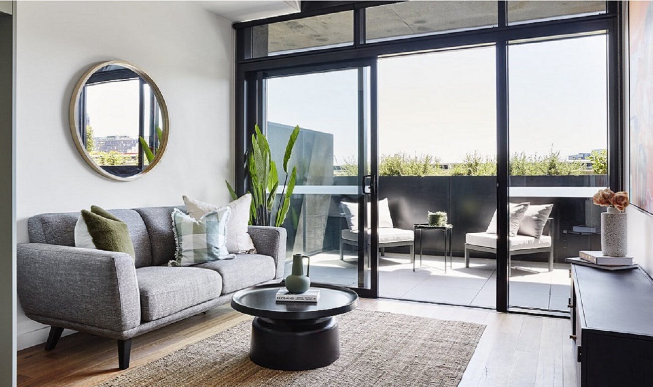 Image of modern living room in an apartment. It features high ceilings, floor-to-ceiling windows and a sliding door to a balcony. The living room has a grey couch, round black coffee table, tan coloured rug and round mirror on the wall.