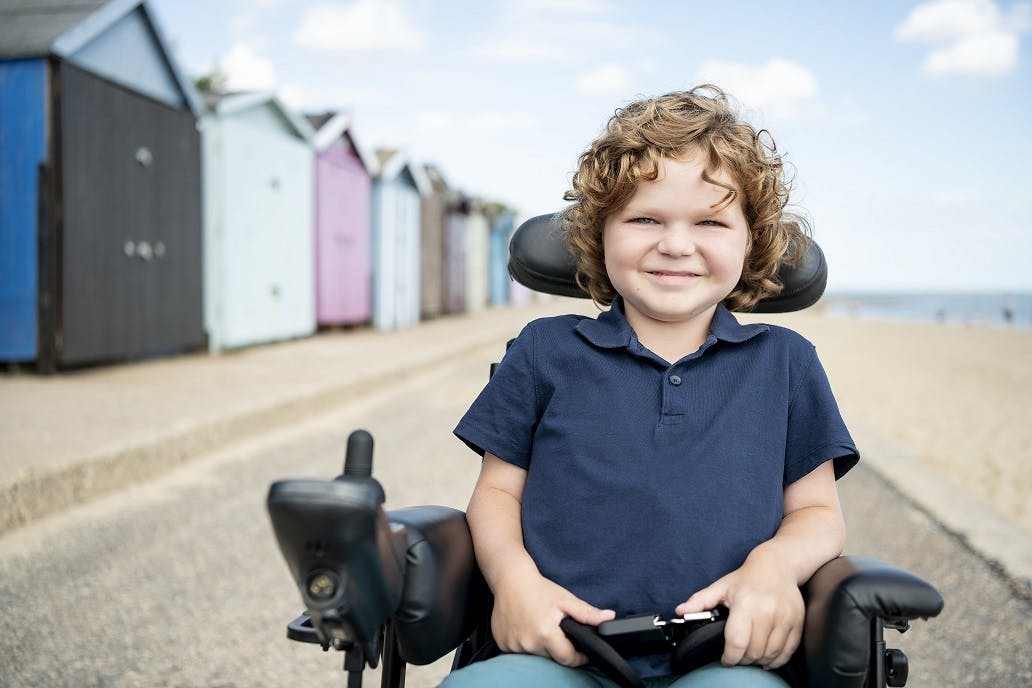 Image a young boy with light-coloured curly hair, smiling at the camera. He is a wheelchair user. There is a bright blue sky, beach and colourful bathing boxes behind him.